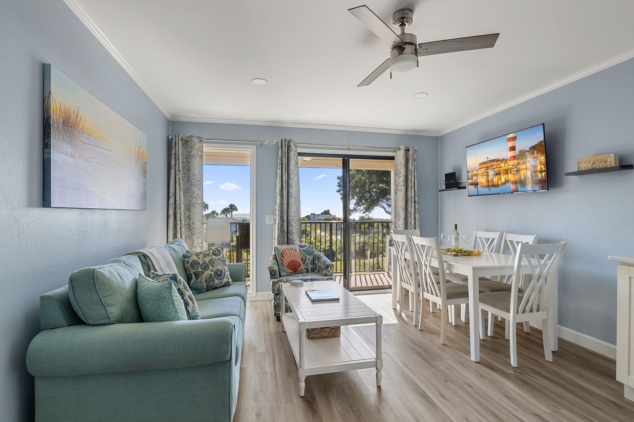 A modern living room with a sofa, coffee table, dining table, and chairs. There is a ceiling fan, a TV, and a painting on the wall.