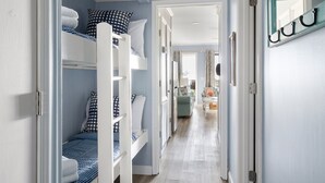 Un pasillo estrecho con literas a la izquierda, una puerta con el letrero 'uu' a la derecha y una vista parcial de una sala de estar al fondo.
