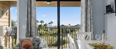 Ein Balkon mit Blick auf Palmen, ein Esstisch mit Obstplatte und Weingläsern sowie eine Sitzecke mit einem Kissen in Muschelform.