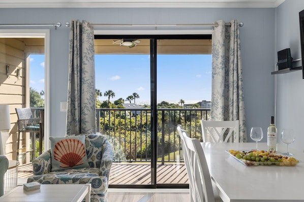 A balcony with a view of palm trees, a dining table with a fruit platter and wine glasses, and a sofa with a shell pillow.