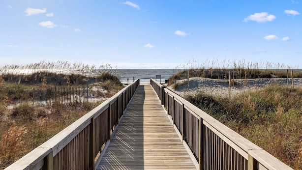 Un paseo marítimo de madera que conduce a la playa, rodeado de dunas de arena y hierbas.
