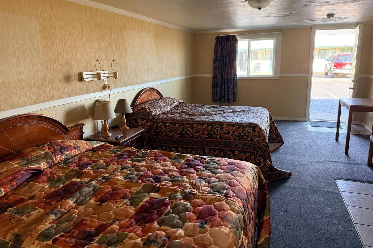 Ein Hotelzimmer mit zwei Betten, einem fensterbelaubten Fenster, einer zur Außenseite führenden Tür und einer Deckenleuchte.