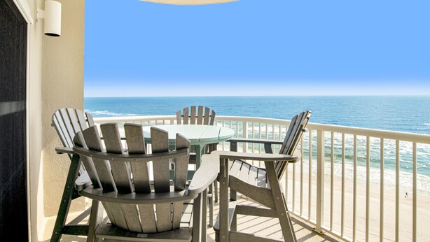 A balcony with two Adirondack chairs and a table, overlooking a beach with the ocean and a clear sky.