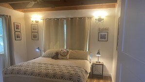 Quarto com cama, mesa de cabeceira, abajur, ventoinha no teto e janela com cortinas.