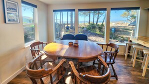 A dining area with a round wooden table and six chairs, large windows with a view of trees and a beach, and framed pictures on the wall.