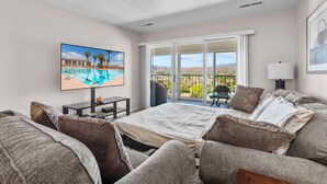 A hotel room with a large bed, a TV displaying a pool view, a balcony with a view of the mountains, and a dining area with a table and chairs. - Las Palmas 326, Resort pool and clubhouse, Private balcony (Saint George)