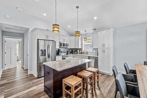 A modern kitchen with white cabinets, stainless steel appliances, and a central island with stools. - Oceanside, Private Pool, Hot Tub, Close to Lighthouse, Community Amenities, VOH23, The Strandhaus (Corolla)