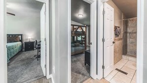 A bedroom with a bed, bedside table, and lamp; a bathroom with a shower and tiled walls; and a hallway with a door leading to another room. - Horseshoe Canyon | Cozy 3BR Cabin • Pet Friendly (Ruidoso)
