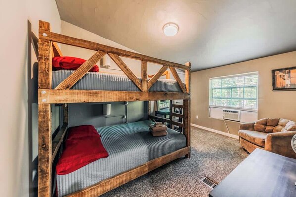 A bunk bed with a wooden frame, a red pillow, and a grey mattress. There is a small table with a lamp, a window with a view, and a couch in the room.