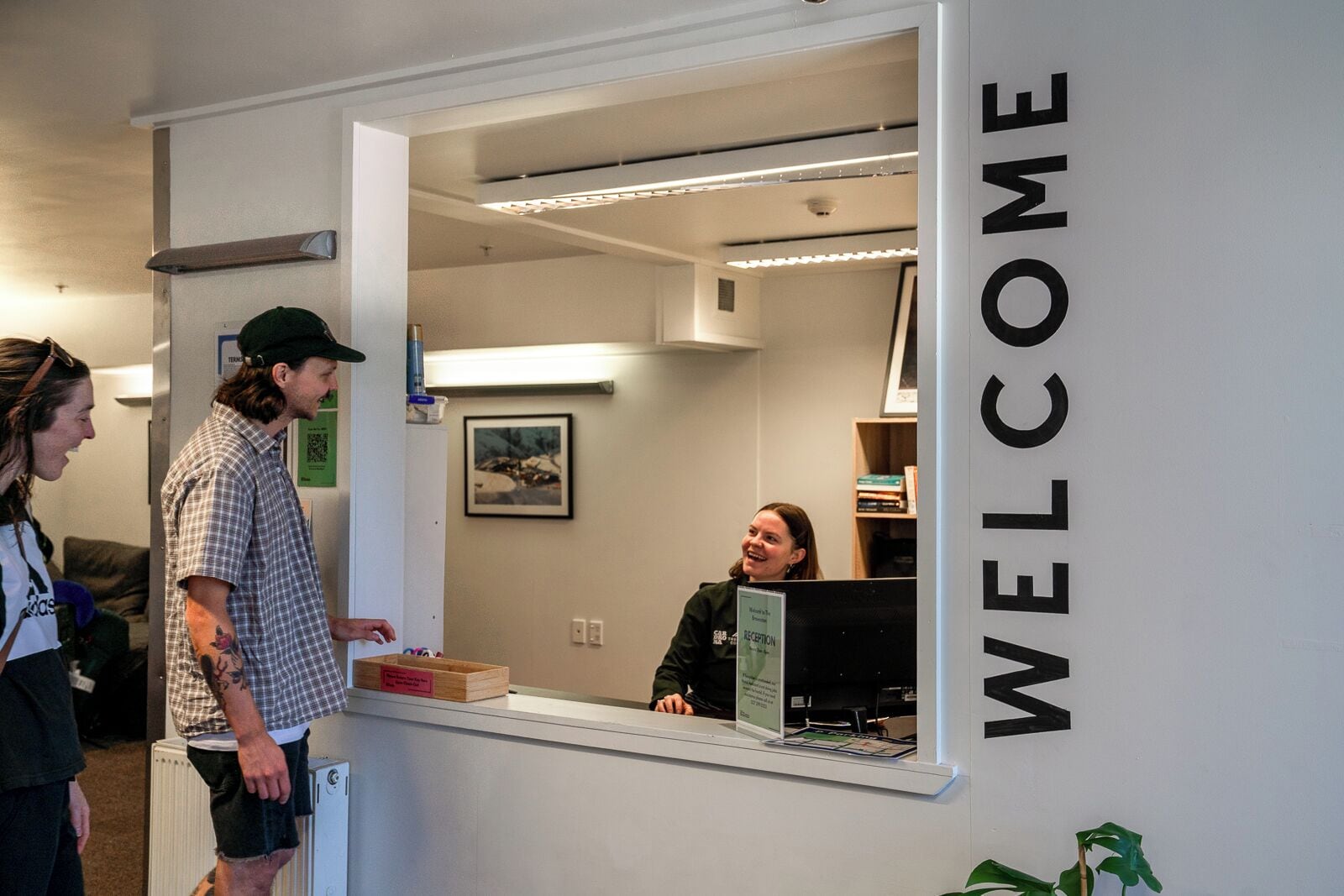 Un comptoir d’accueil avec une pancarte « BIENVENUE », un tableau encadré accroché au mur et une personne assise derrière le comptoir.
