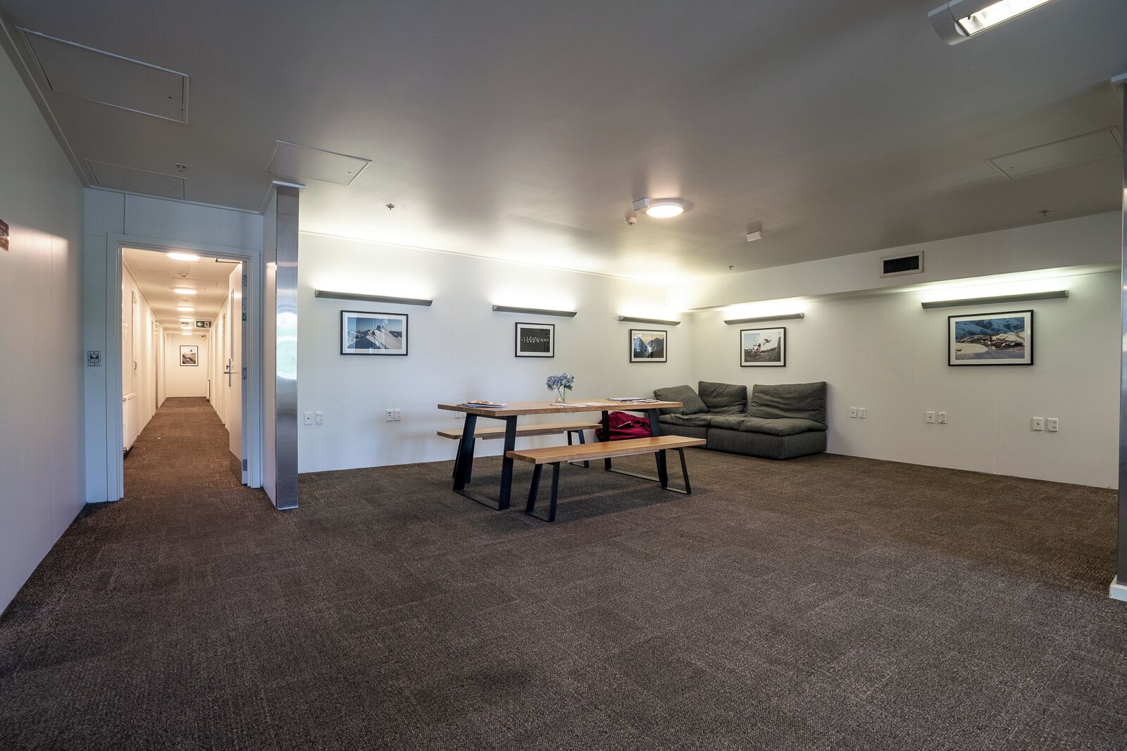 Una habitación amplia con piso alfombrado, una mesa de madera, un sofá gris y cuadros enmarcados en las paredes.
