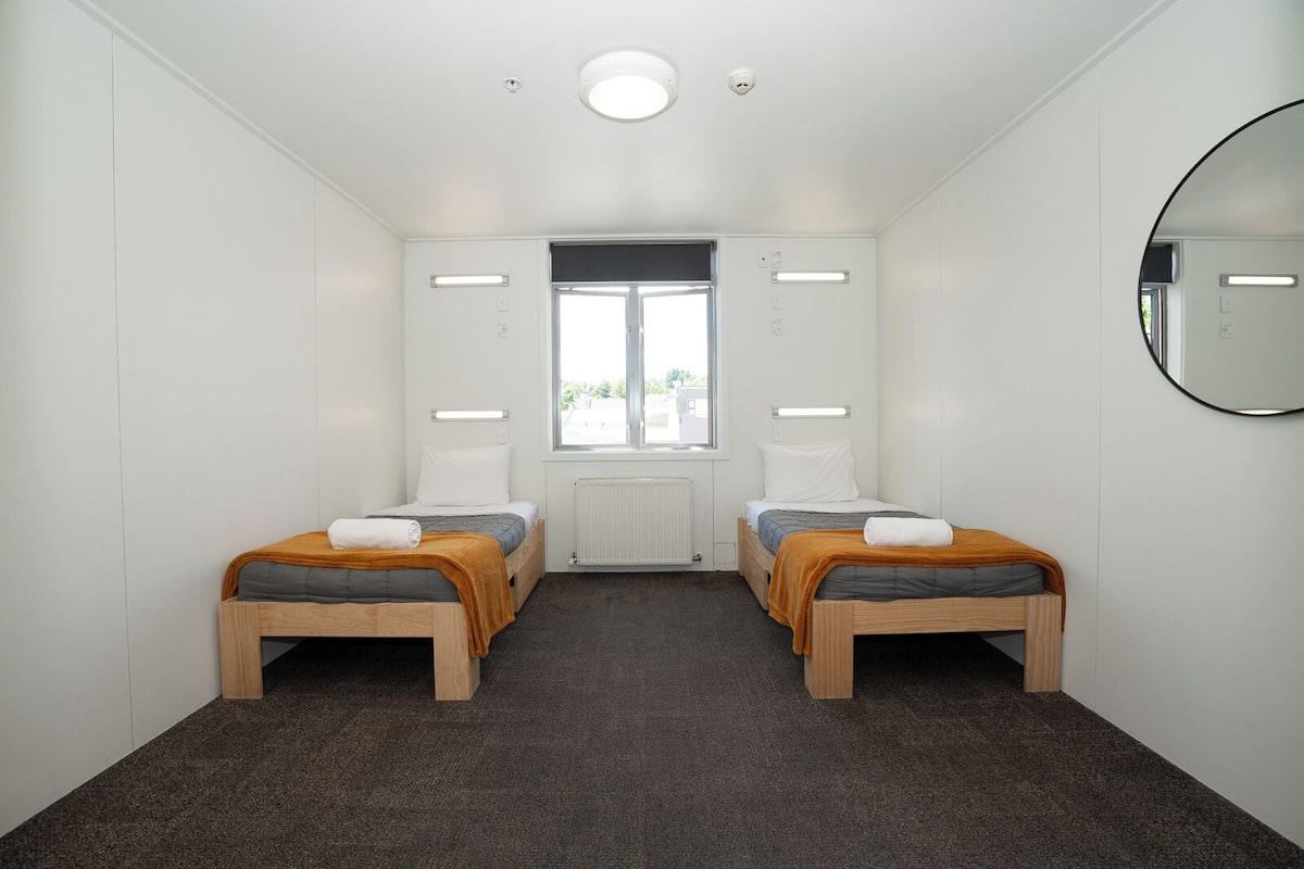 A small, modern room with two single beds, a large window, and a mirror on the wall.