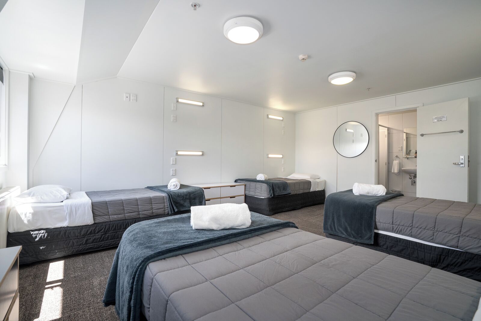 A hotel room with three beds, a desk, a mirror, and a bathroom visible through an open door.
