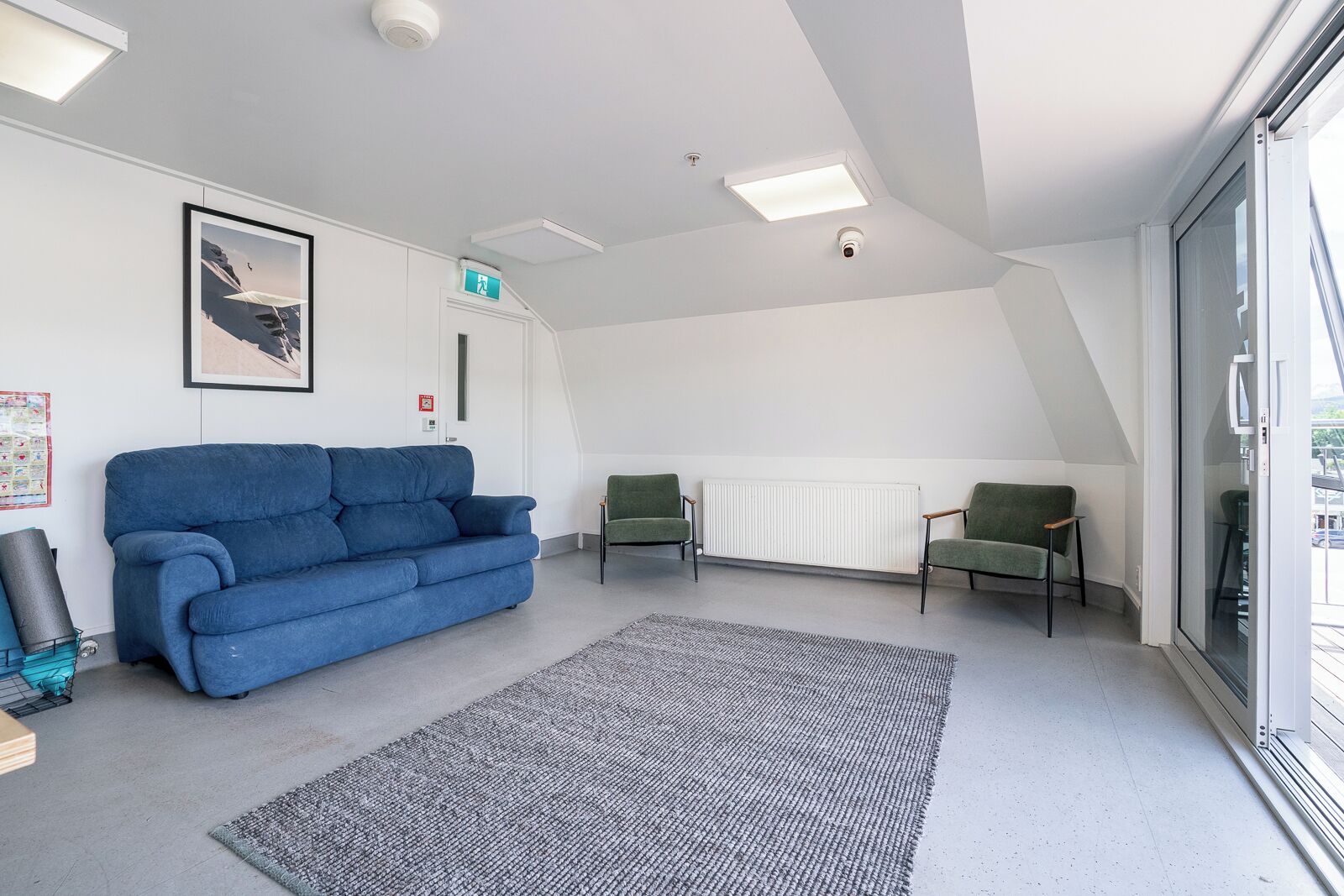 Een moderne woonkamer met een blauwe bank, twee groene fauteuils, een grijze vloerkleed en een witte radiator.