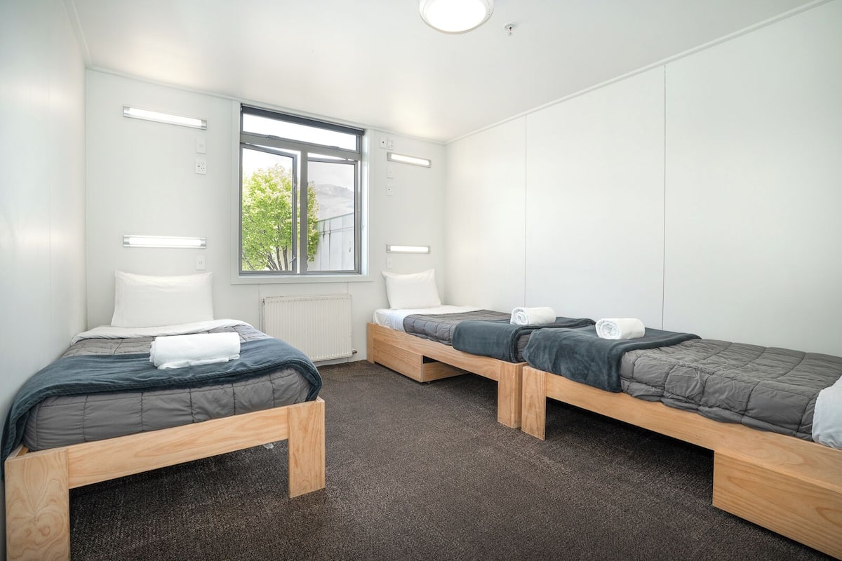 A dormitory room with three wooden bunk beds, white bedding, and a large window providing natural light.