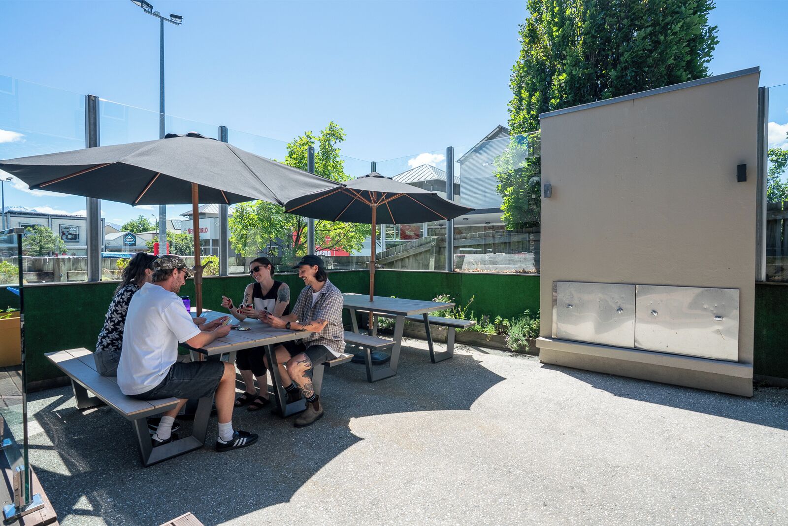Un grupo de personas sentadas en una mesa al aire libre con sombrillas, una pared verde y un edificio al fondo.