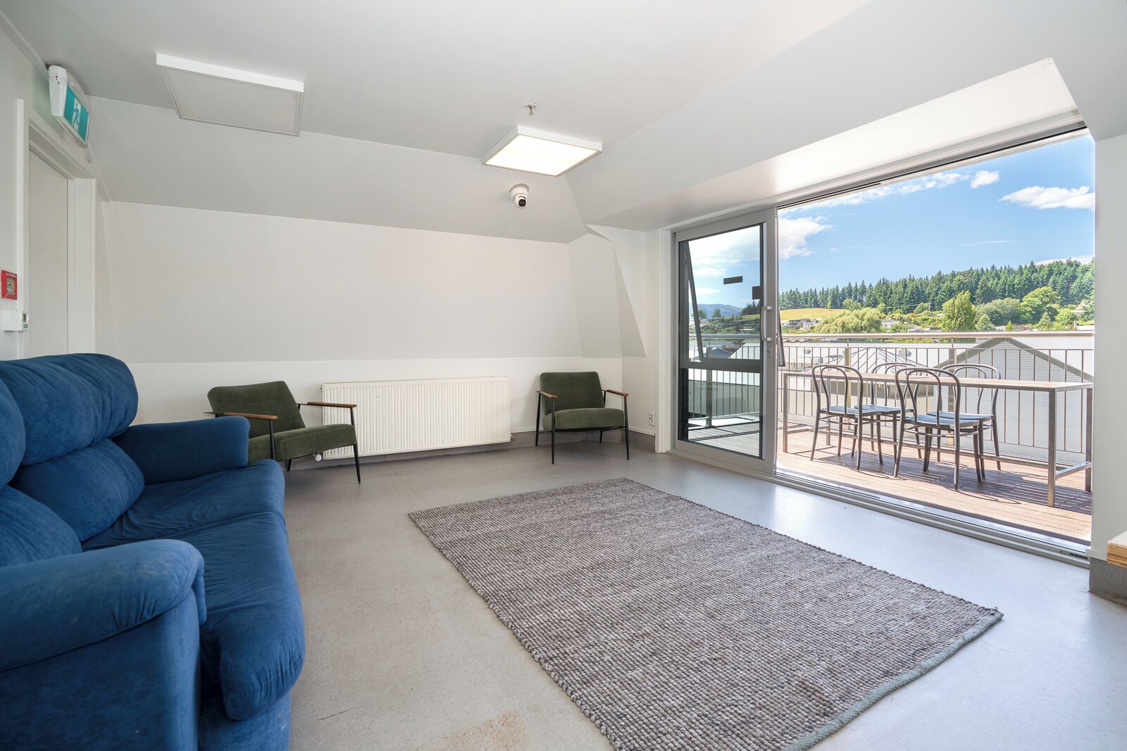 Un salon moderne avec un canapé bleu, deux fauteuils verts, un tapis gris et un balcon aménagé avec du mobilier d’extérieur.