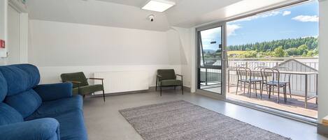 Un salon moderne avec un canapé bleu, deux fauteuils verts, un tapis gris et un balcon aménagé avec du mobilier d’extérieur.