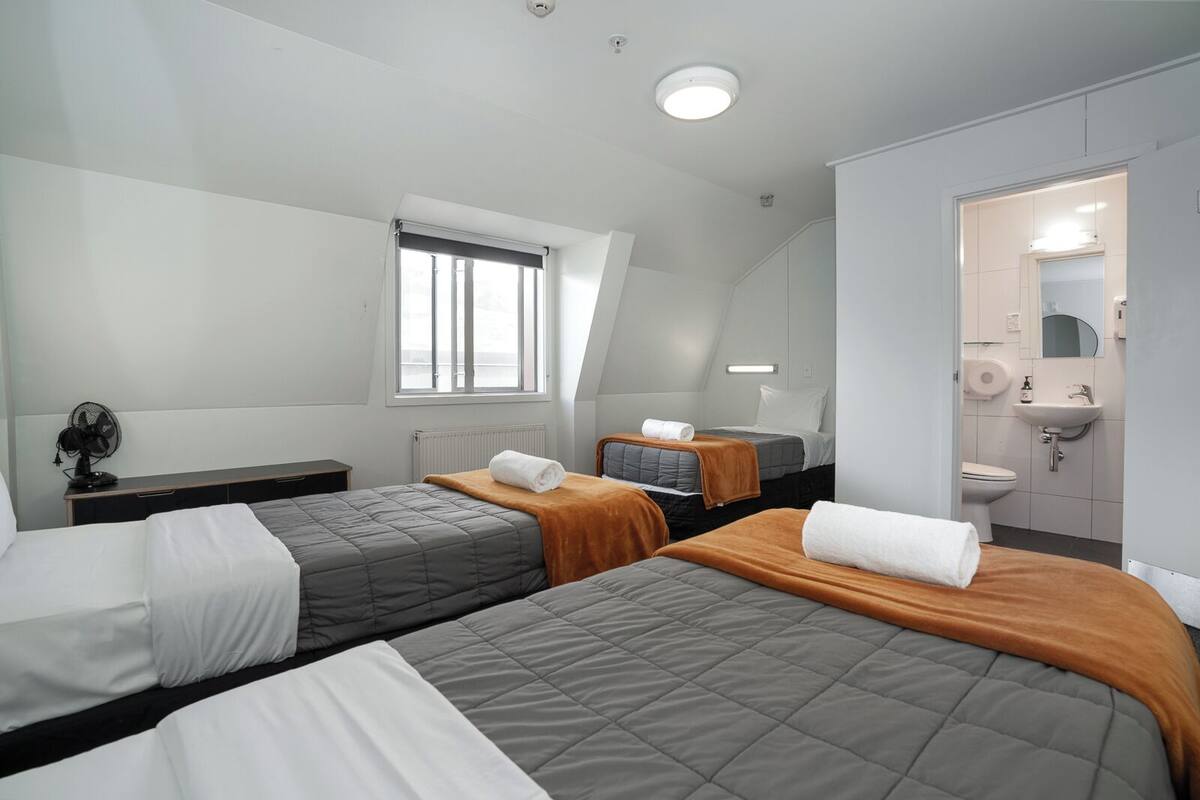 A hotel room with two beds, a window, a radiator, a fan, a sink, and a mirror.