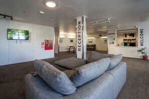 A reception area with a grey sofa, a TV on the wall, and a 'WELCOME' sign. - The Brownston Hostel (Wanaka)