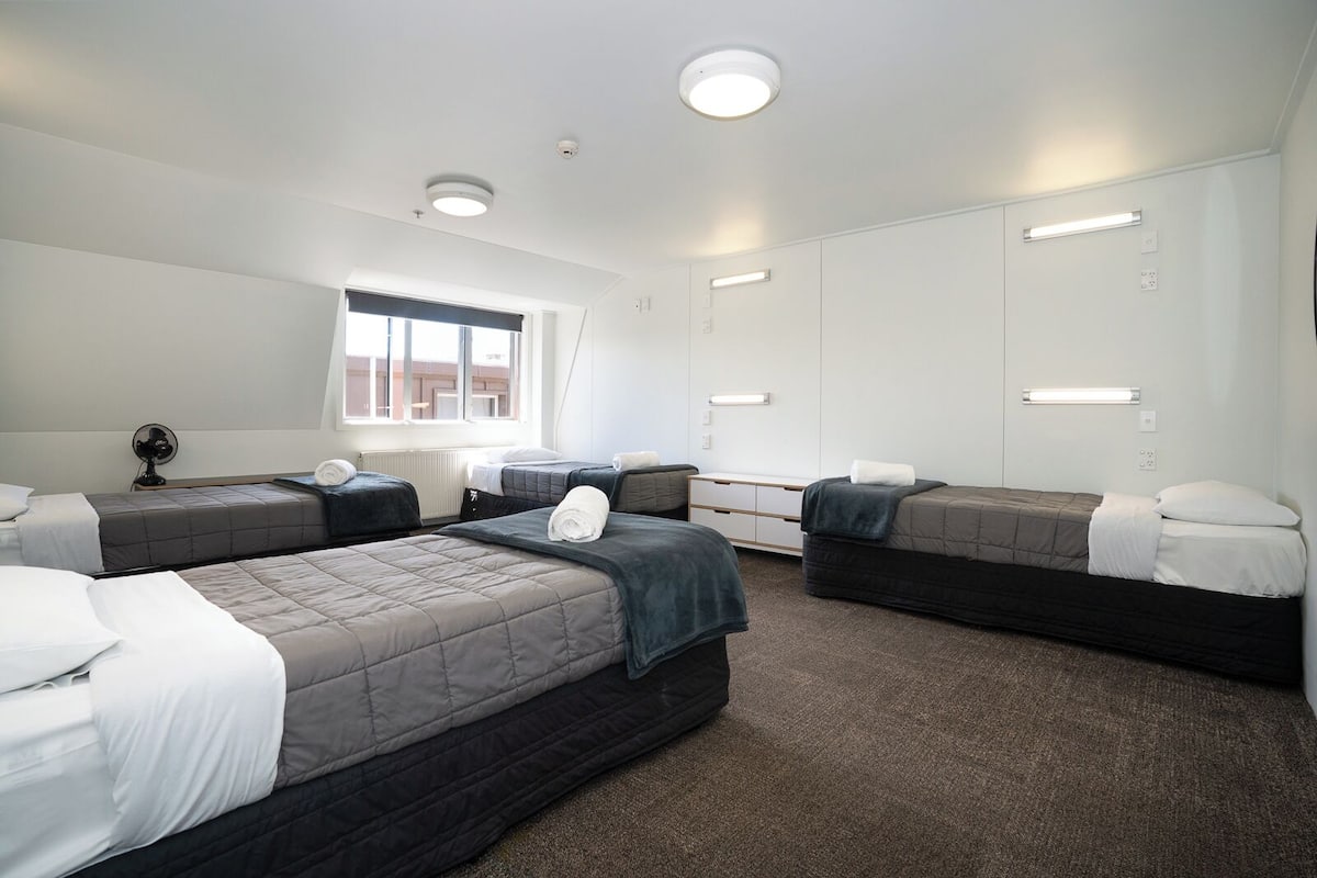 A hotel room with four beds arranged in a U-shape, a large window with blinds, a ceiling fan, and recessed lighting.