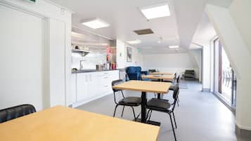 Cocina moderna con armarios blancos y un sofá azul, zona de comedor con mesas de madera y sillas negras, y ventanas amplias que dejan entrar luz natural.