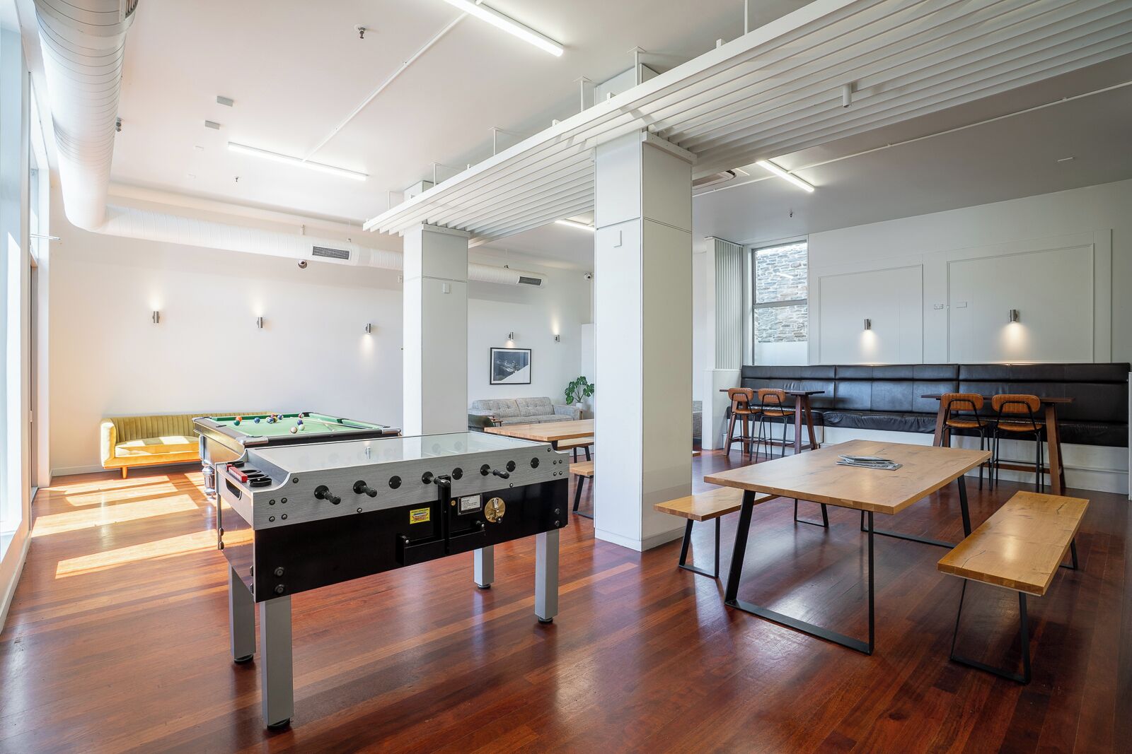 A modern indoor space with a foosball table, wooden tables, and benches, featuring a clean, minimalist design with white walls and wooden flooring.