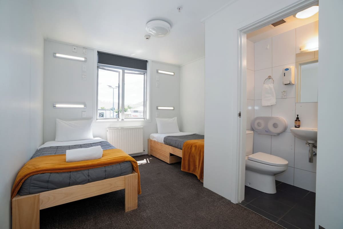 A modern hotel room with two single beds, a large window, a bathroom with a toilet and sink, and a ceiling light.