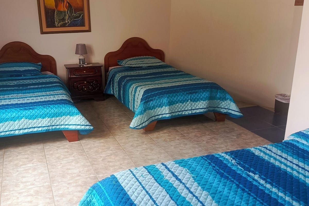A room with two beds, each with a blue quilted bedspread, a nightstand with a lamp, and a painting on the wall.