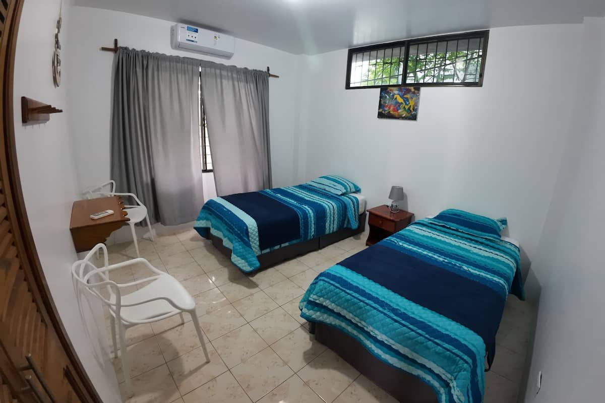A bedroom with two beds, a desk, a chair, an air conditioner, and a window with a view of trees.