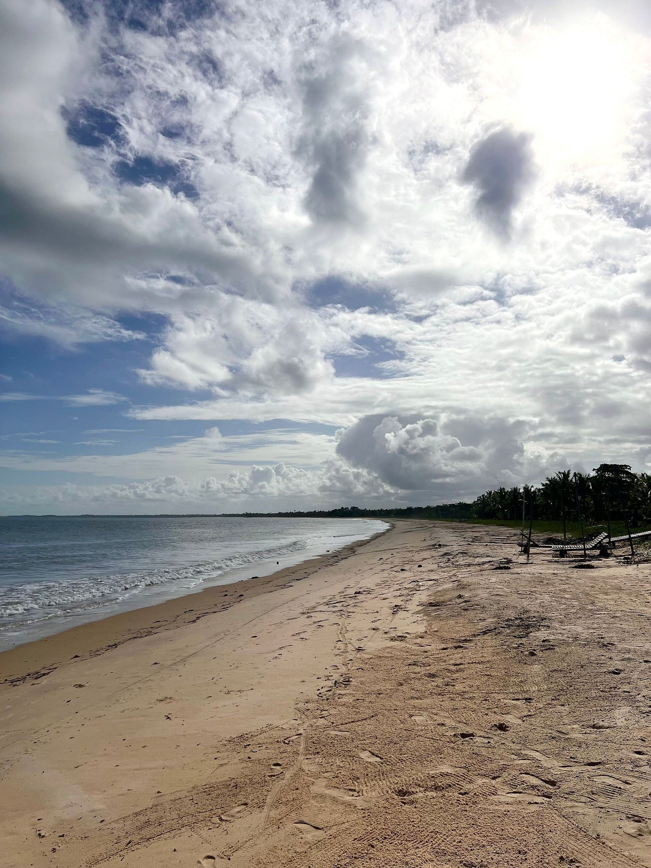 A beach with a clear sky and clouds, sandy shore, and palm trees in the distance.