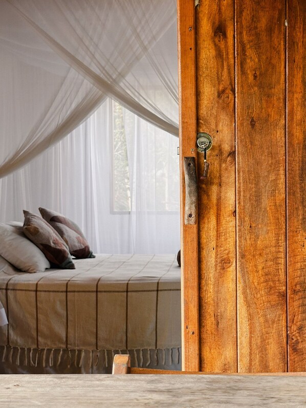 A bedroom with a large wooden door, a bed with a beige bedspread, and a sheer white canopy. - Casa da Mata Corumbau (Prado)