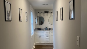 A hallway with wooden flooring, white walls, and framed artwork. There are two round mirrors and a sink with a marble countertop.