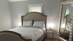 A bedroom with a bed, a ceiling fan, a window, a bedside table, and a large mirror.