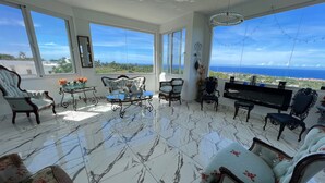 Amplia habitación con un gran ventanal que ofrece vista al océano, piso de mármol y muebles clásicos que incluyen un sofá, sillas y una mesa.