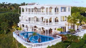 Un edificio blanco de varios pisos con piscina, rodeado de abundante vegetación.