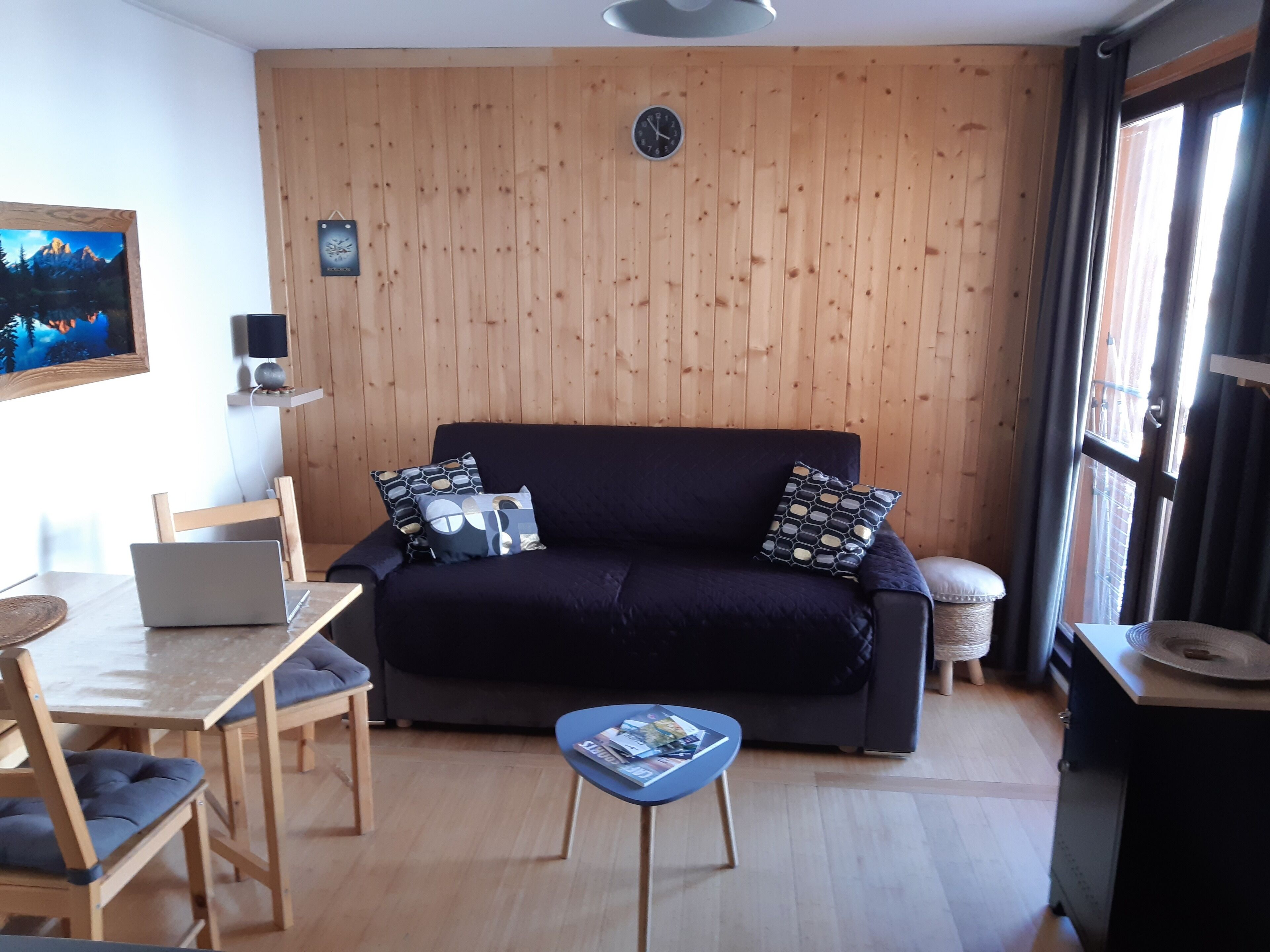 Un soggiorno accogliente con un divano blu scuro, pareti in legno, un tavolo da pranzo in legno con sedie, un portatile e un piccolo tavolino blu con riviste.