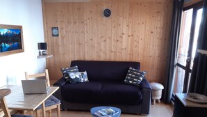 A cozy living room with a dark blue sofa, wooden walls, a wooden dining table with chairs, a laptop, and a small blue side table with magazines. - Studio Cosy Les Deux Alpes Centre (Les Deux Alpes)
