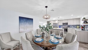 A modern dining area with a round wooden table set for six, surrounded by six grey upholstered chairs. A decorative hanging light fixture is above the table. - Spacious beach home - Sleeps 12 - Steps from dining, with a private heated pool (Marco Island)