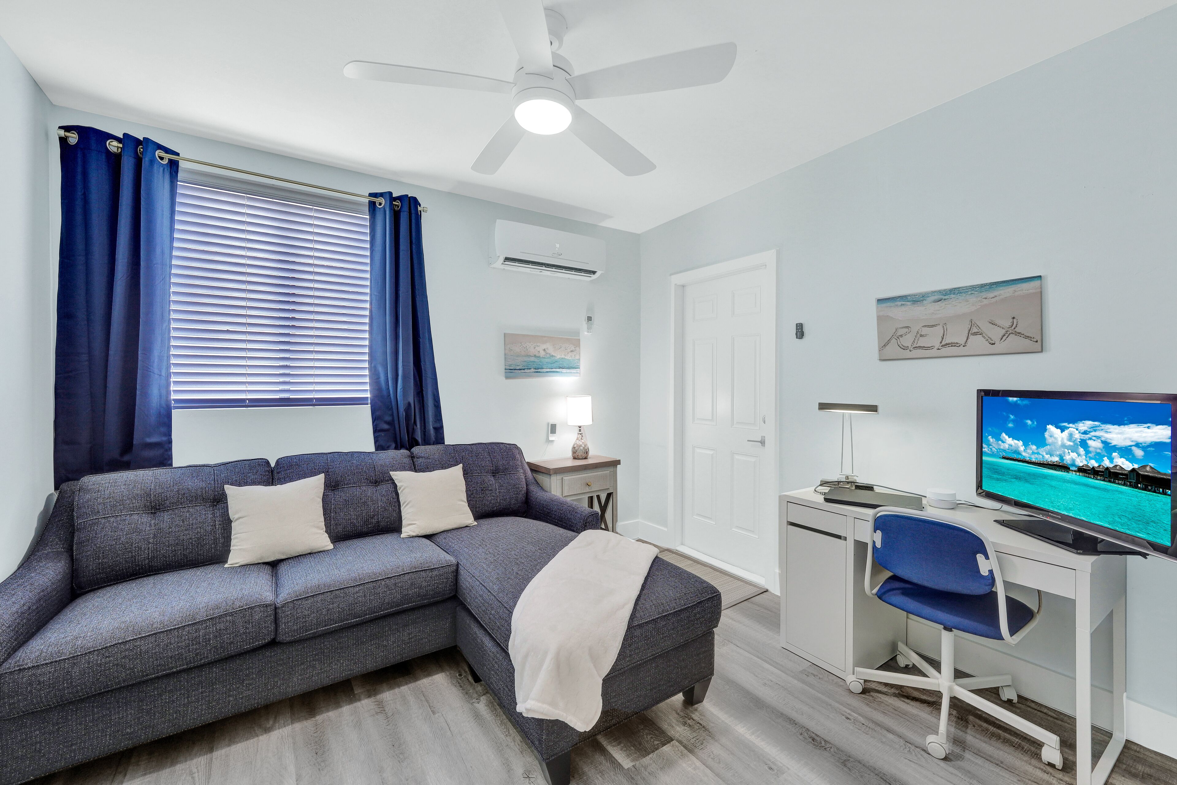 A modern living room with a grey sectional sofa, a white desk with a computer and TV, and a ceiling fan.
