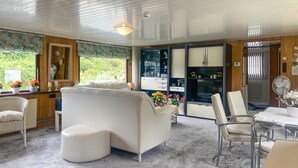 Un salon chaleureux avec un canapé blanc, une table à manger et un téléviseur. On y trouve des chaises en osier et une petite table ornée de fleurs.