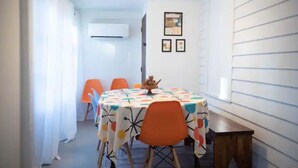 Un comedor con una mesa redonda cubierta con un mantel colorido, rodeada de sillas naranjas. A la derecha, hay una pequeña mesa de madera.