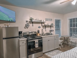 A compact kitchen with a stove, oven, and refrigerator. There is a television mounted above the stove, a small dining table with chairs, and a window with blinds.