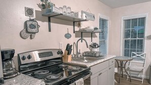 Una cocina con estufa negra, un fregadero y una cafetera. Hay elementos decorativos en la pared y una ventana con persianas.
