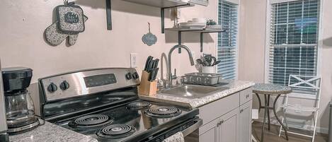 Una cocina con estufa negra, un fregadero y una cafetera. Hay elementos decorativos en la pared y una ventana con persianas.