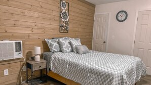 Un dormitorio con un panel de pared de madera, una cama con ropa de cama estampada, una mesita de noche con lámpara, un equipo de aire acondicionado y un reloj en la pared.