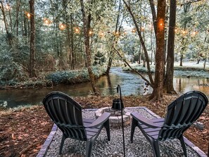 Un espace extérieur paisible, aménagé avec deux chaises et une table, entouré d’arbres et bordé d’un ruisseau.
