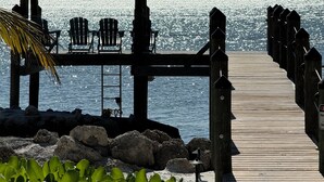 Un muelle de madera que lleva a una zona de playa con una estructura de techo de paja, sillas y vistas al mar.