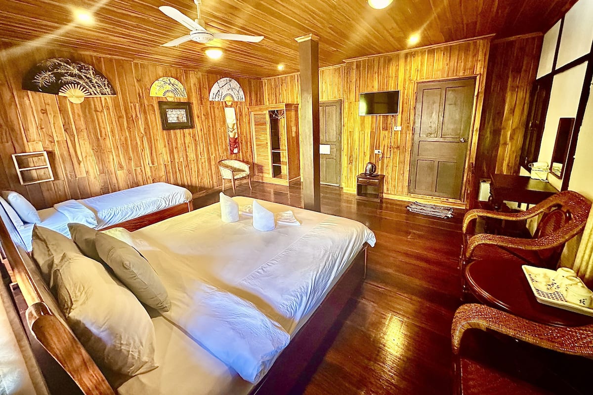 A wooden-themed room with a bed, two chairs, a TV, and a ceiling fan.