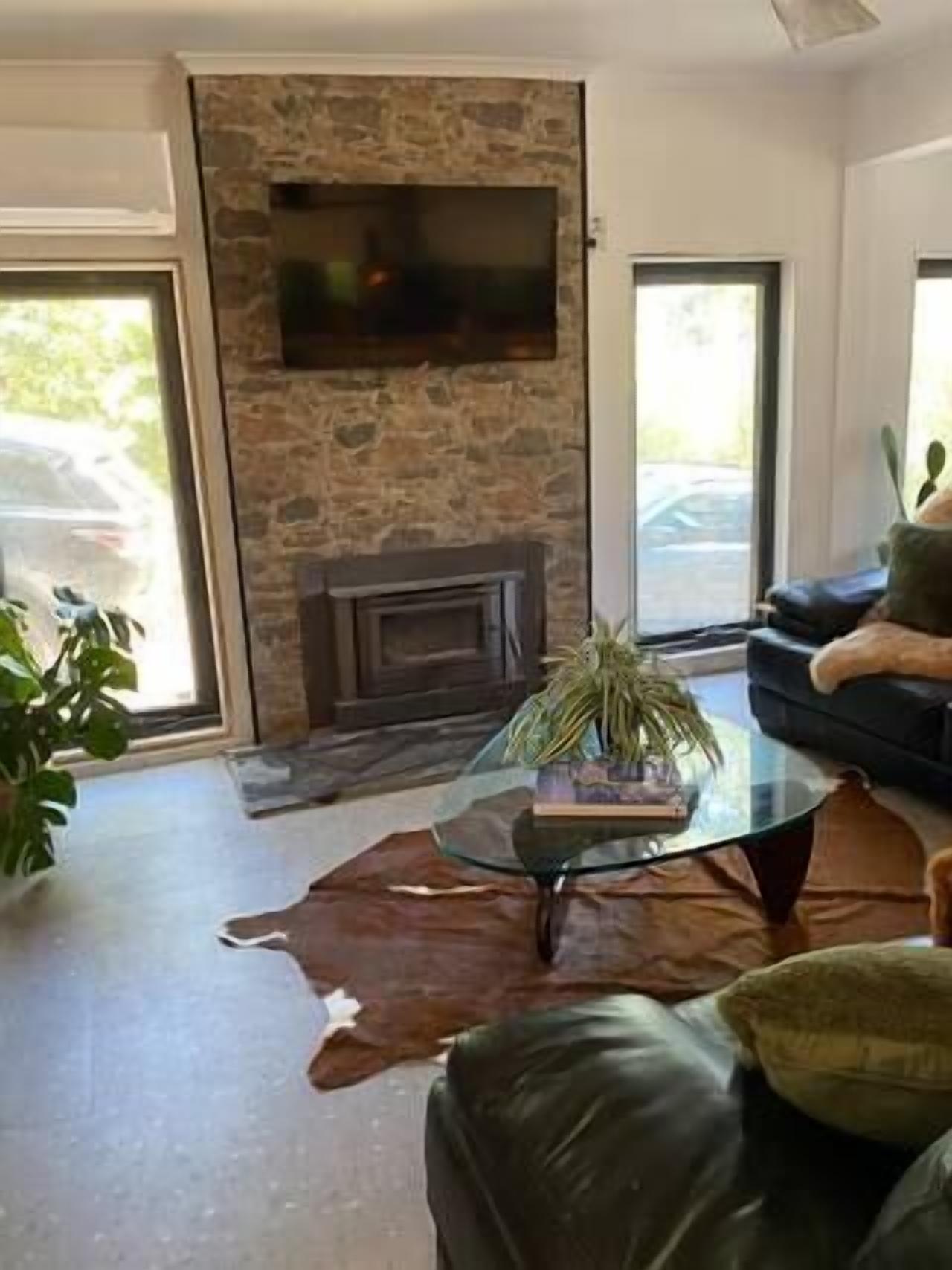 A modern living room with a stone fireplace, a glass coffee table with a plant, a leather sofa, and a large window.